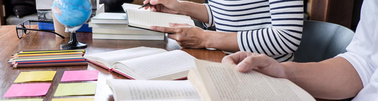Two adults sitting at a table with open books, sticky notes, pencils on desk