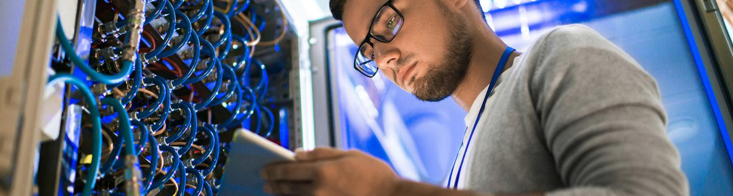 Low angle portrait of young man using digital tablet standing by server cabinet while working with supercomputer in blue light