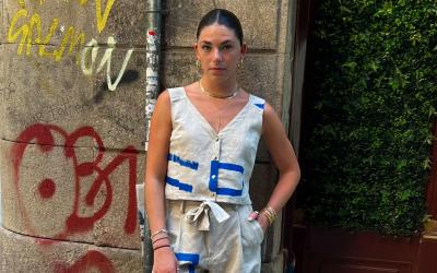 Person standing on a city street, dressed in a contemporary outfit featuring white and blue designs, accessorized with a white bag and sneakers. Graffiti is visible on the wall beside them.
