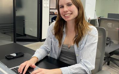 Girl sitting at a desk typing on a computer while smiling for a picture. 