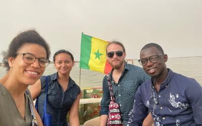 Four people sitting together on a boat with the flag of Senegal visible in the background. They are smiling and appear to be enjoying their time on the water.